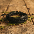 Pulsera multicapa cuero negro entrelazado ajustable sobre madera con hojas verdes